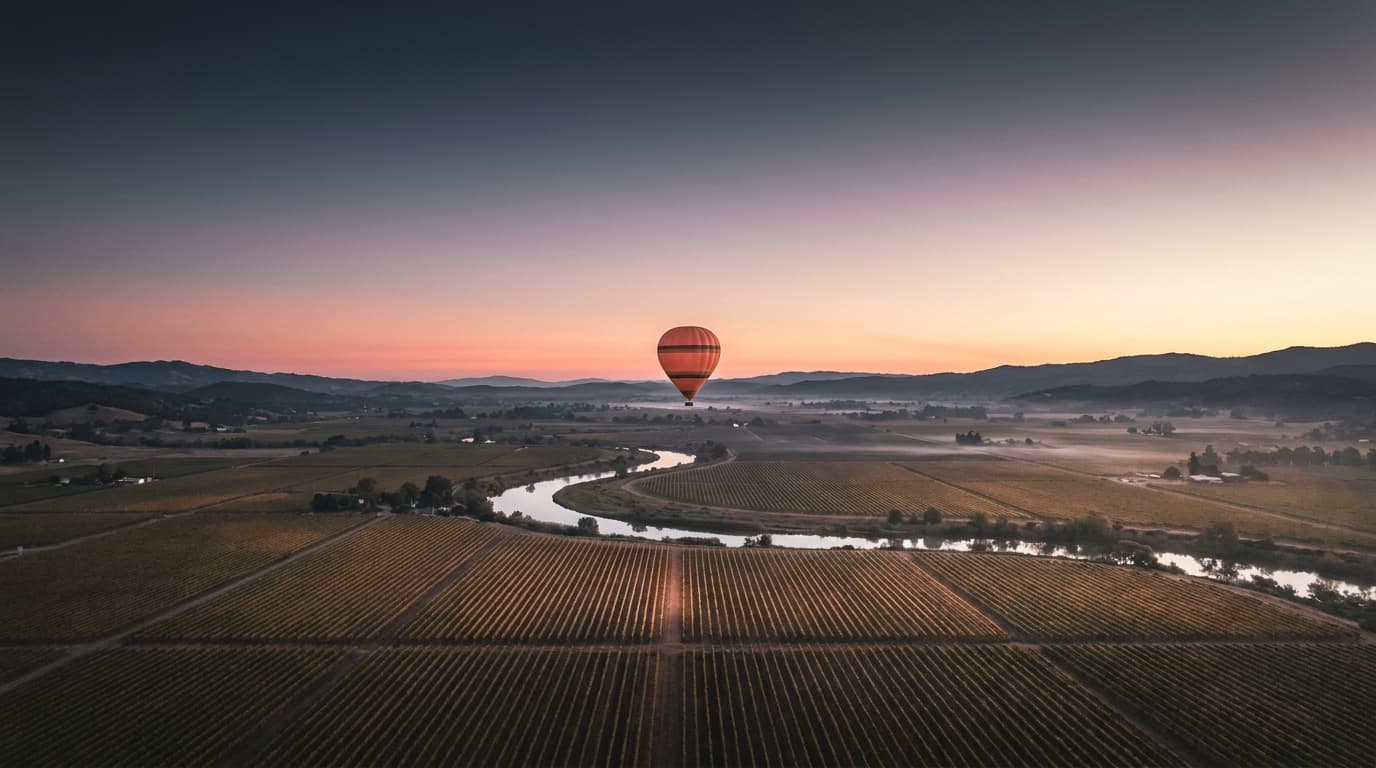 A single Drift and Dawn hot-air balloon aloft at altitude over the Sonoma vineyards and a silver curving river at dawn; the sky reads from deep navy at the upper edge through lavender and coral to pale gold along the horizon, faint ground mist in the low places