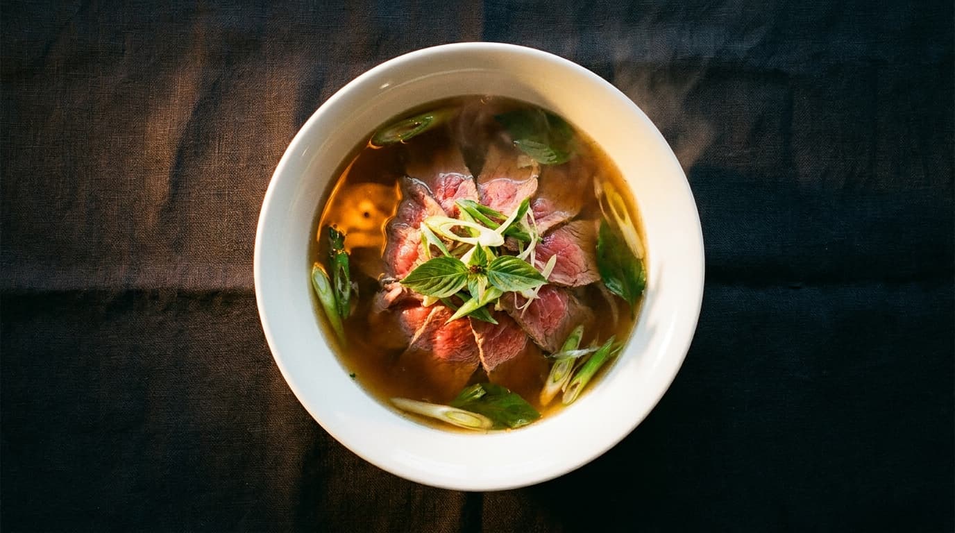 Edge-to-edge overhead photograph of a single bowl of phở on dark linen