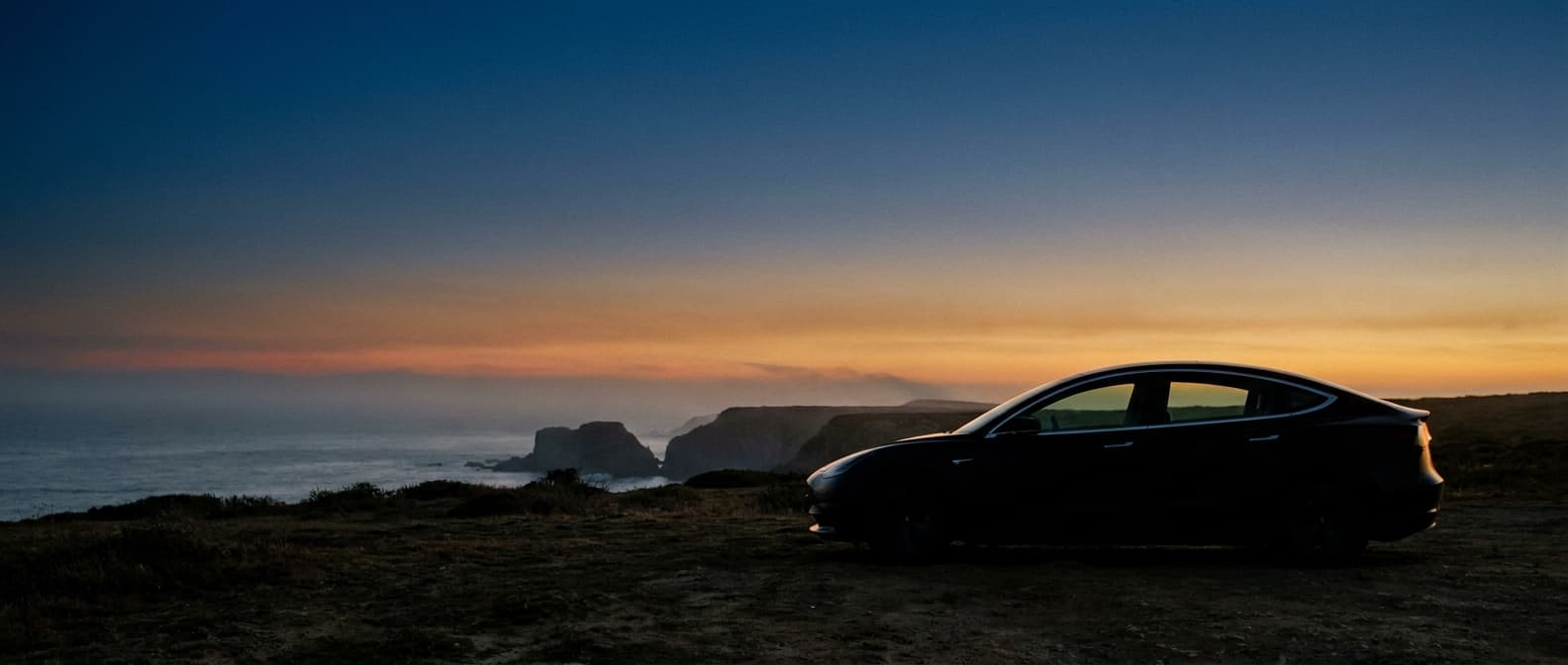 Anode hero: a premium electric sedan in three-quarter front view at dusk against a coastal landscape.