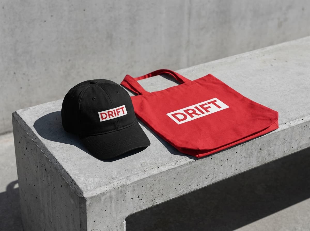 A street still: a Drift cap and tote on a concrete bench with hard daylight.