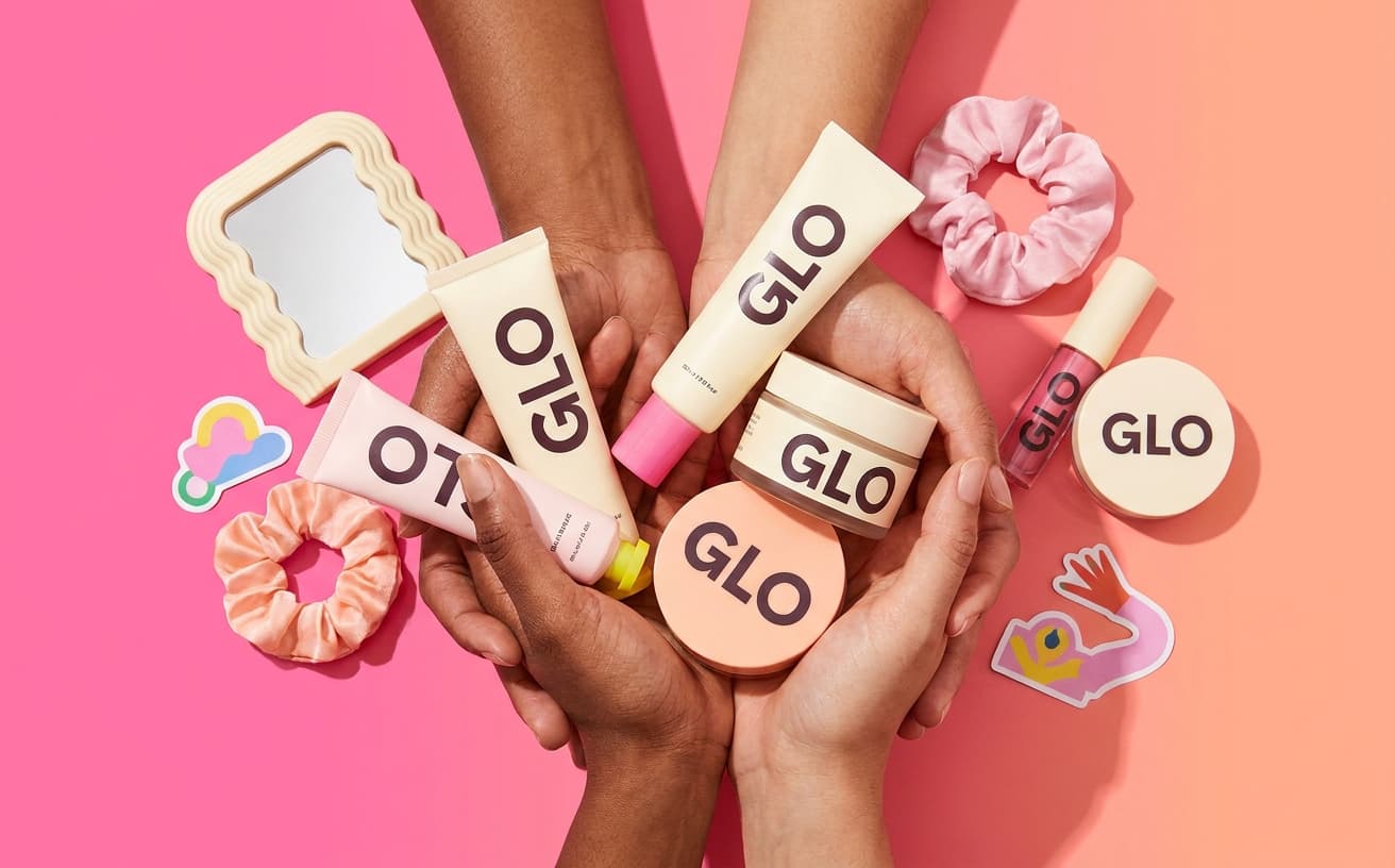 A close-up of hands holding multiple Glo products on a pink and peach gradient background with playful props.