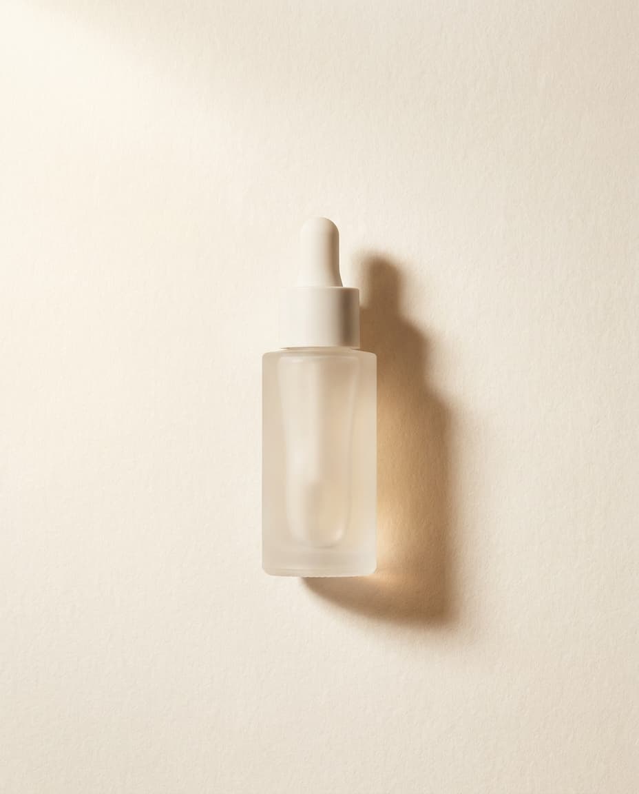 A slim ceramic-stoppered bottle of pale serum on a stone shelf, side-lit, with a single linen edge in the frame.